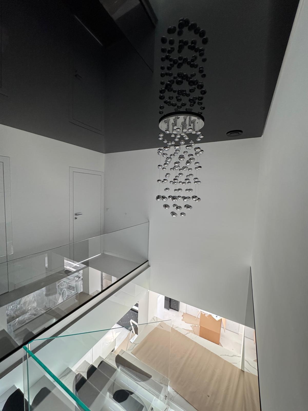 Nowoczesne wnętrze z czarnym, lustrzanym sufitem napinanym i designerskim żyrandolem. Widok z góry na szklane balustrady i częściowo zabezpieczone podłoże.