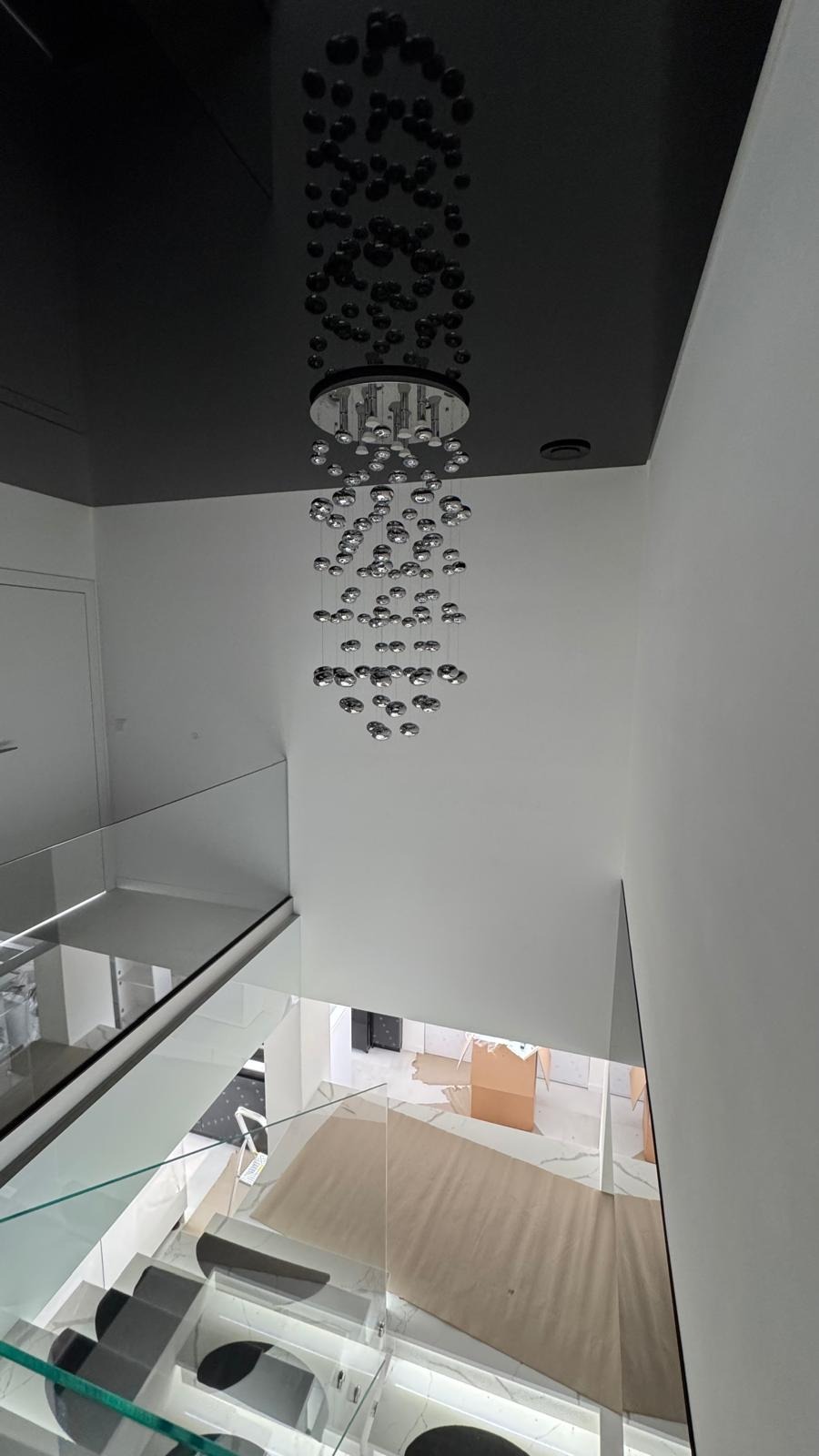 Nowoczesny, wysoki hol z czarnym sufitem i designerską lampą z chromowanych kul. Widok z góry na szklane balustrady i marmurowe schody. Minimalistyczny wystrój.