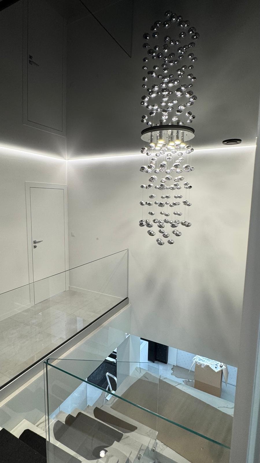 Nowoczesne wnętrze z efektownym, srebrnym żyrandolem kaskadowym, szklanymi balustradami i białymi ścianami z oświetleniem LED. Widok z góry na schody.