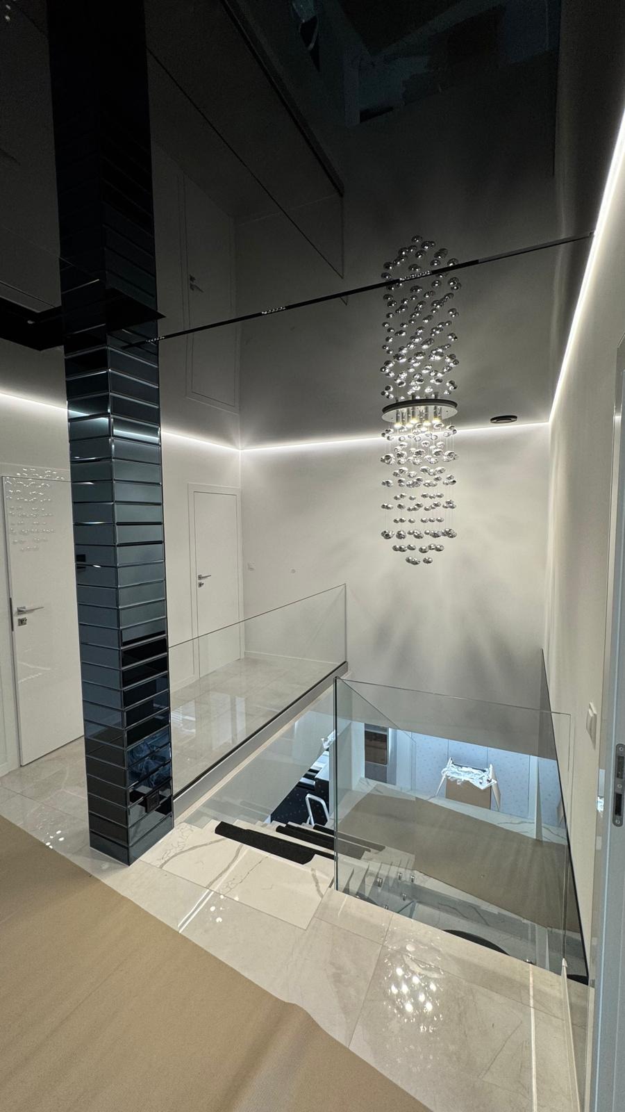 Nowoczesne wnętrze z lustrzanym sufitem, kolumną z płytek i szklanymi balustradami schodów. Żyrandol z kul.