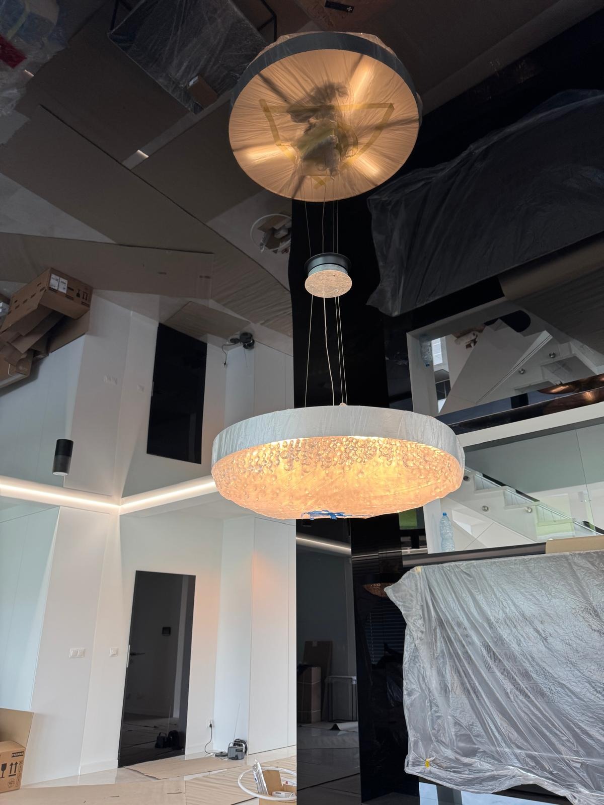 Nowoczesny, jasny żyrandol z ozdobnymi kryształkami w przestronnym wnętrzu podczas wykańczania. Widoczne zabezpieczenia folią malarską i kartony.