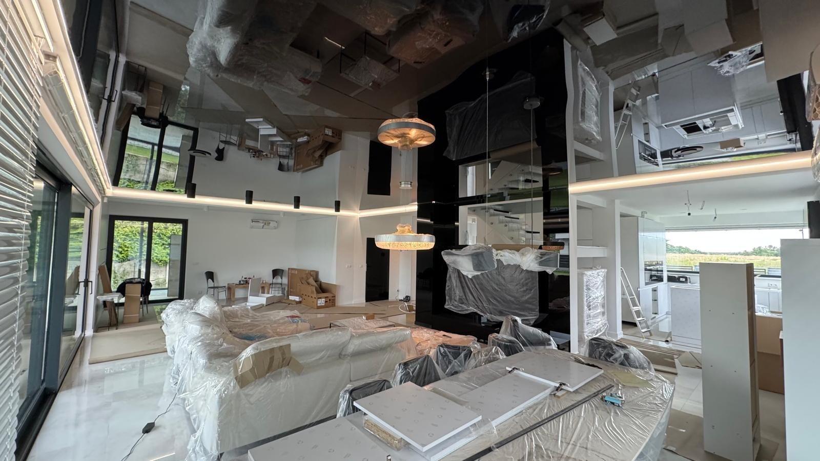 Wnętrze domu w trakcie wykończenia: meble zabezpieczone folią, widoczne elementy oświetlenia i fragment kuchni. Sufit lustrzany. Prace wykończeniowe.