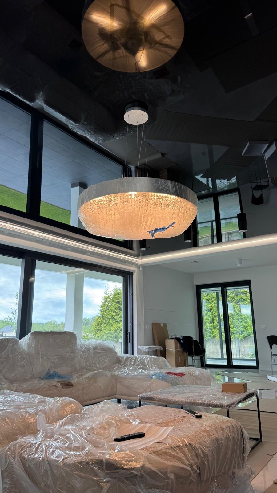 Nowoczesny salon z lśniącym, czarnym sufitem napinanym i designerską lampą. Meble zabezpieczone folią ochronną. Duże okna z widokiem na zieleń.
