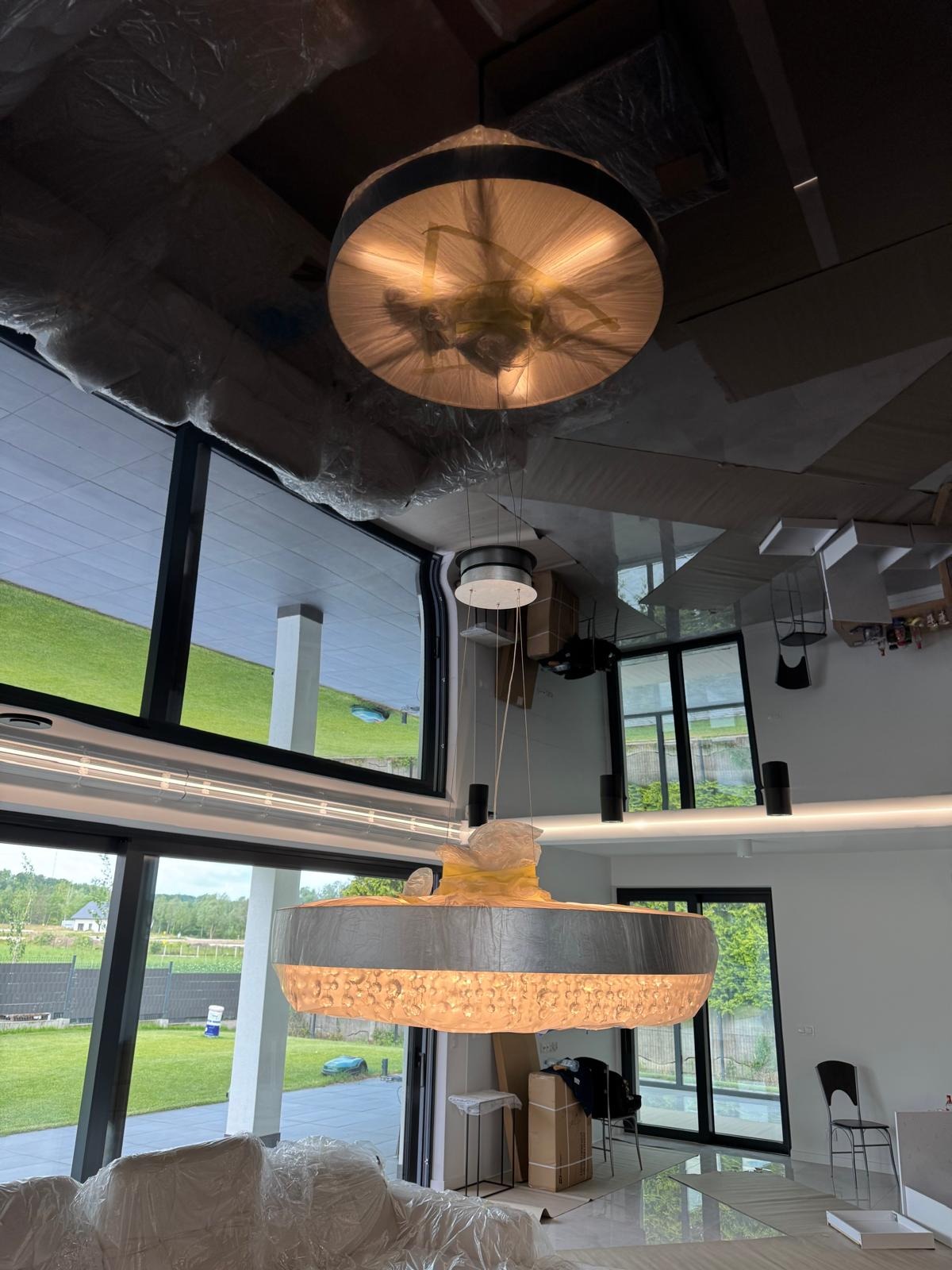 Nowoczesny salon w trakcie wykańczania: duże okna z widokiem na zewnątrz, dwa okrągłe, ozdobne żyrandole, meble zabezpieczone folią malarską.