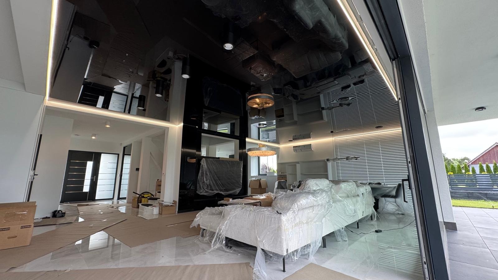 Nowoczesny salon w trakcie wykańczania: czarny, lustrzany sufit z oświetleniem LED, marmurowe podłogi, meble zabezpieczone folią. Widoczne prace wykończeniowe.