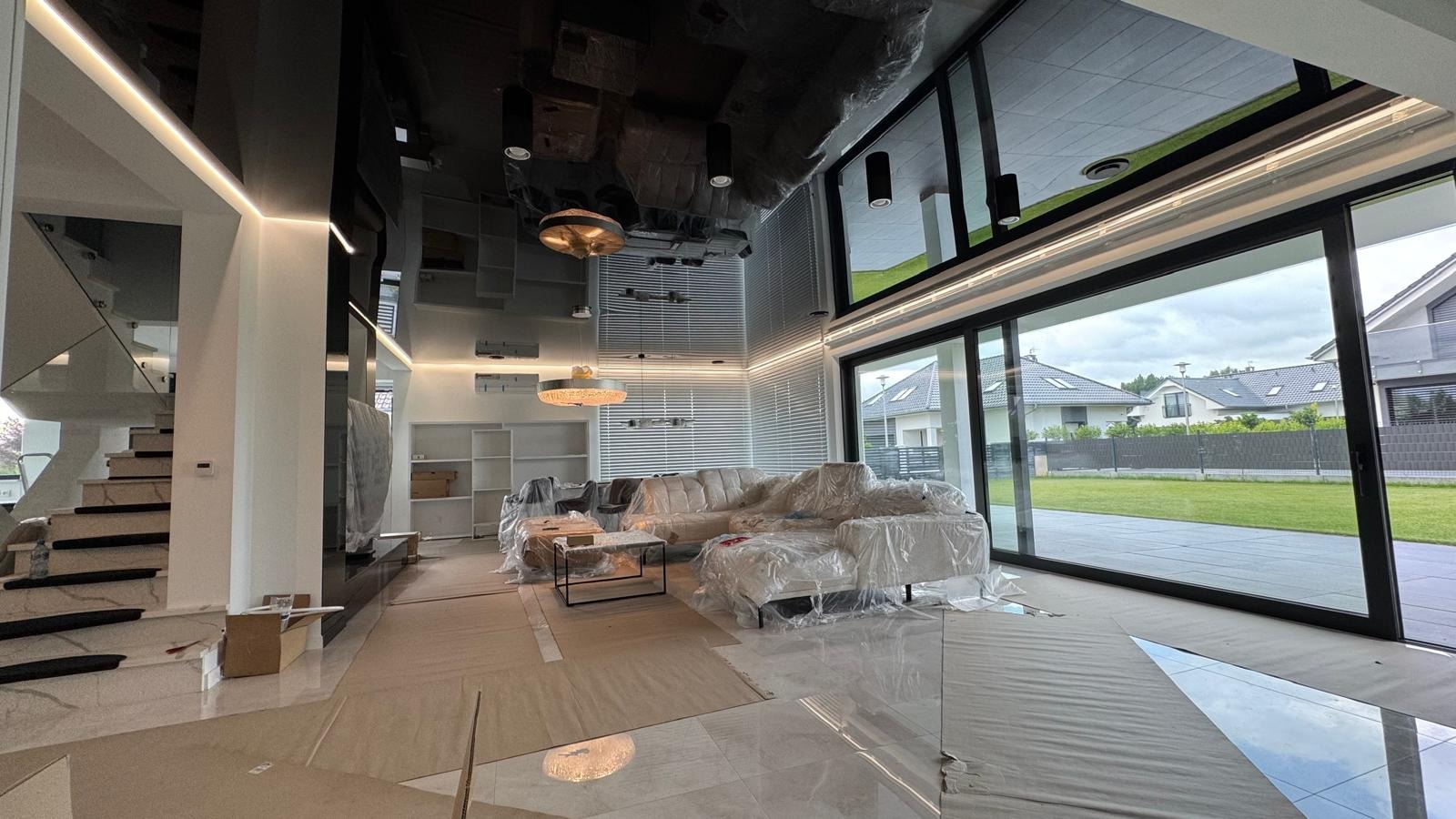 Nowoczesne wnętrze domu w trakcie wykańczania: marmurowe schody, duże okna, błyszczący sufit i podłoga, meble zabezpieczone folią. Widoczne prace wykończeniowe.