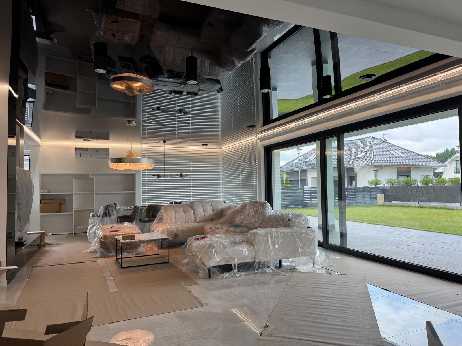 Nowoczesny salon w trakcie zabezpieczania folią przed pracami wykończeniowymi; widoczne meble pod folią, okno tarasowe i połyskliwy sufit.