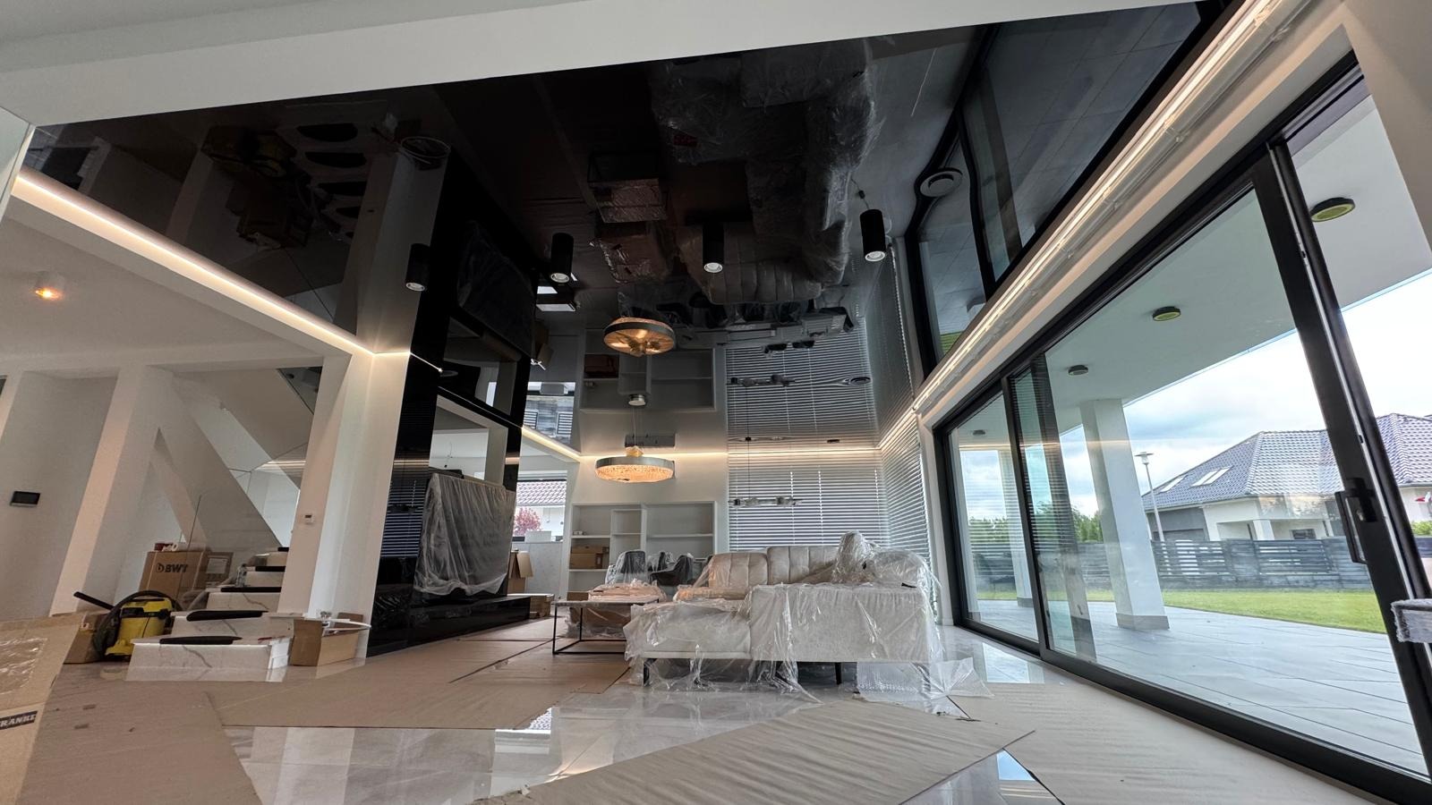 Nowoczesny salon po remoncie, z błyszczącym czarnym sufitem napinanym i dużymi przesuwnymi oknami. Meble zabezpieczone folią, widoczne schody i elementy oświetlenia.