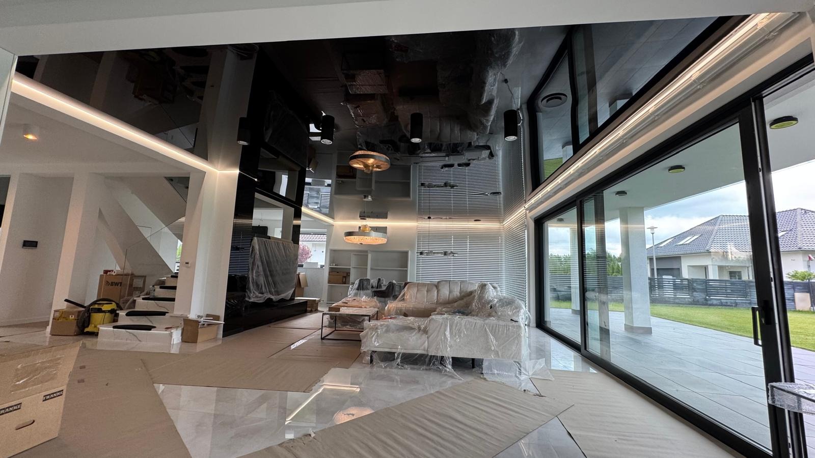Nowoczesny salon z błyszczącym, czarnym sufitem podwieszanym i dużymi przesuwnymi drzwiami tarasowymi. Meble zabezpieczone folią, widoczne prace wykończeniowe.