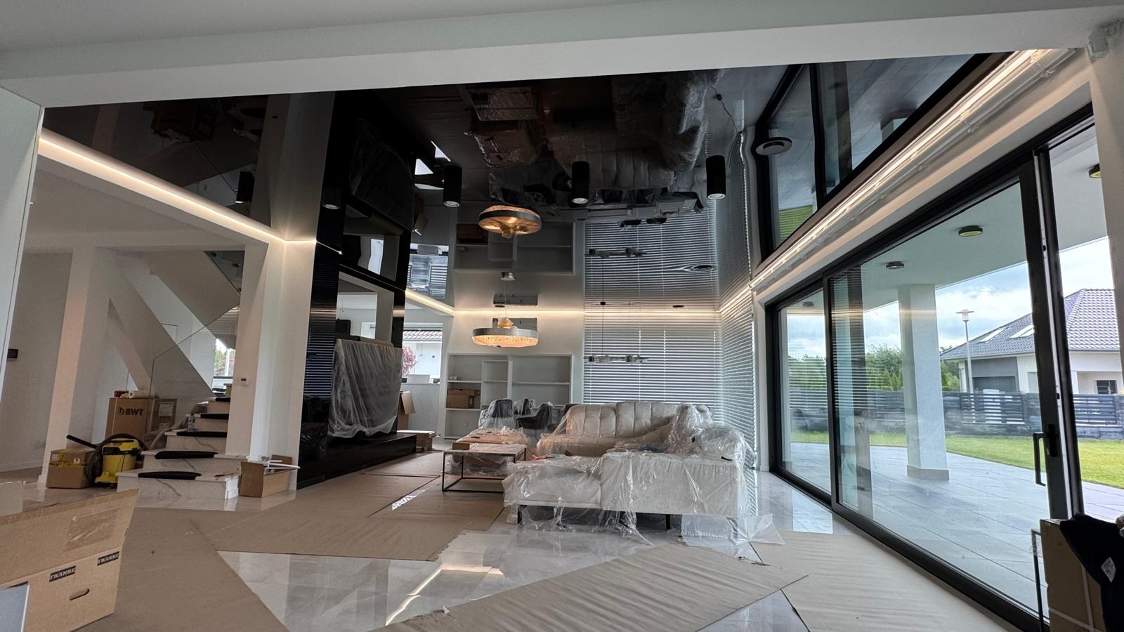 Nowoczesny salon po remoncie z błyszczącym, czarnym sufitem napinanym i dużymi oknami przesuwnymi. Meble zabezpieczone folią malarską, podłoga z płytek.