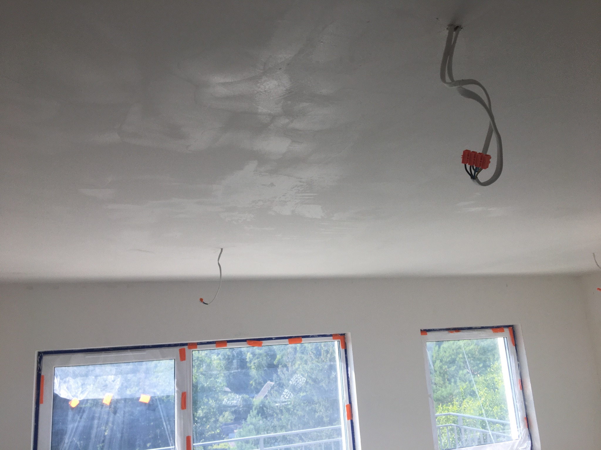 Szpachlowana sufit i ściany w trakcie prac wykończeniowych, zabezpieczone okna folią malarską, wystające przewody elektryczne.