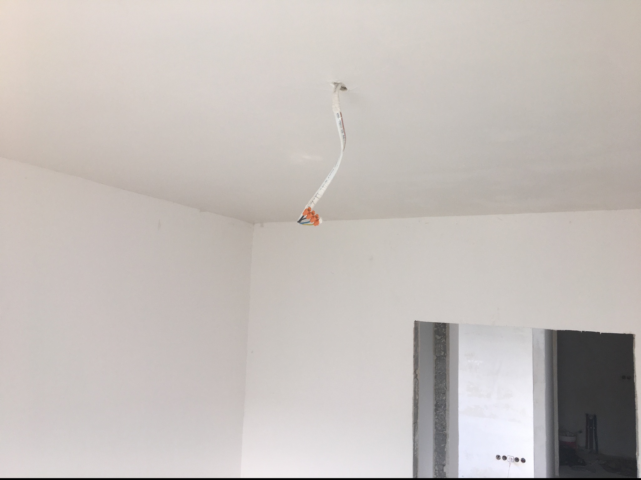 Wnętrze w trakcie prac wykończeniowych: białe ściany, sufit z wystającym przewodem elektrycznym i widocznym otworem, fragment otworu drzwiowego z surowym wykończeniem.