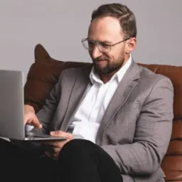 Uśmiechnięty mężczyzna w okularach i marynarce siedzi na skórzanym pufie i pracuje na laptopie Apple, patrząc na ekran.