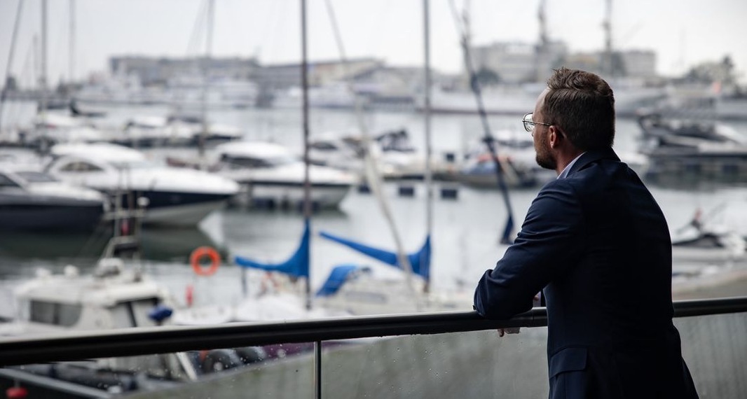 Mężczyzna w garniturze spogląda na zatokę z jachtami, opierając się o szklaną balustradę balkonu.