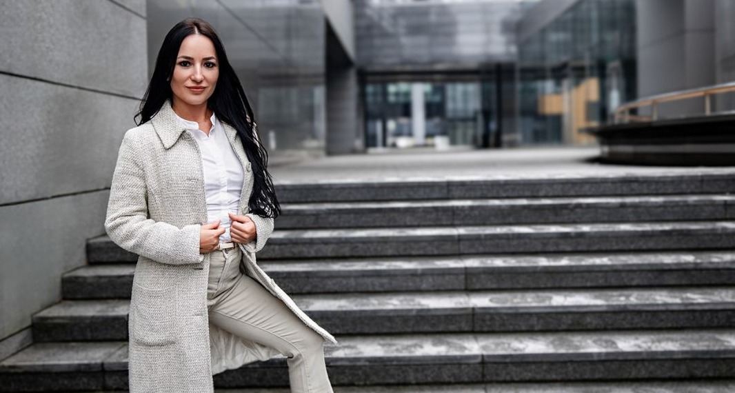 Kobieta w jasnym płaszczu i spodniach pozuje na schodach nowoczesnego budynku biurowego.