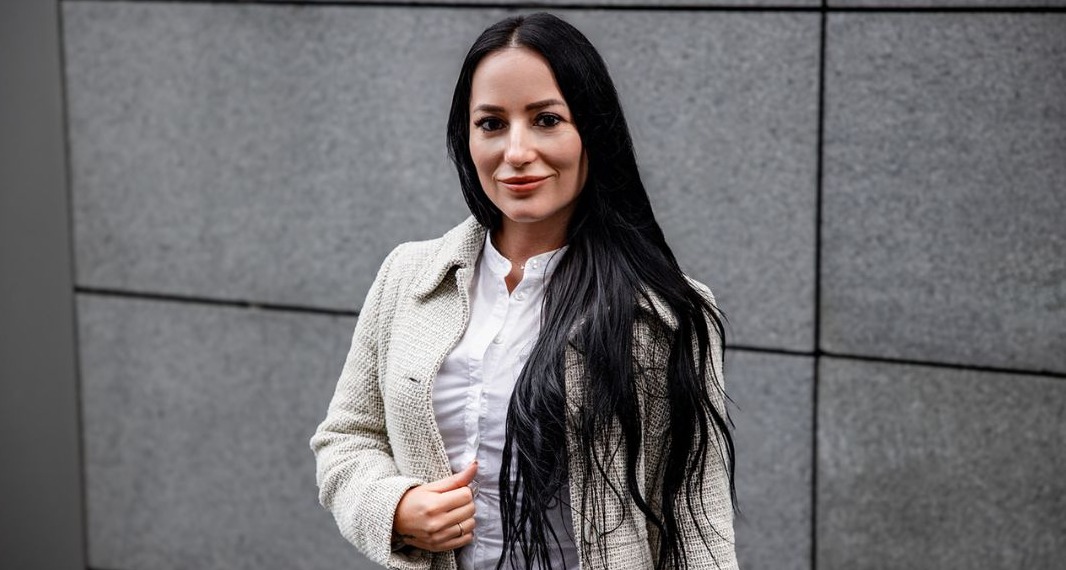 Portret kobiety w jasnym tweedowym płaszczu i białej koszuli na tle szarej ściany z poziomymi liniami, uśmiechająca się, z długimi czarnymi włosami.