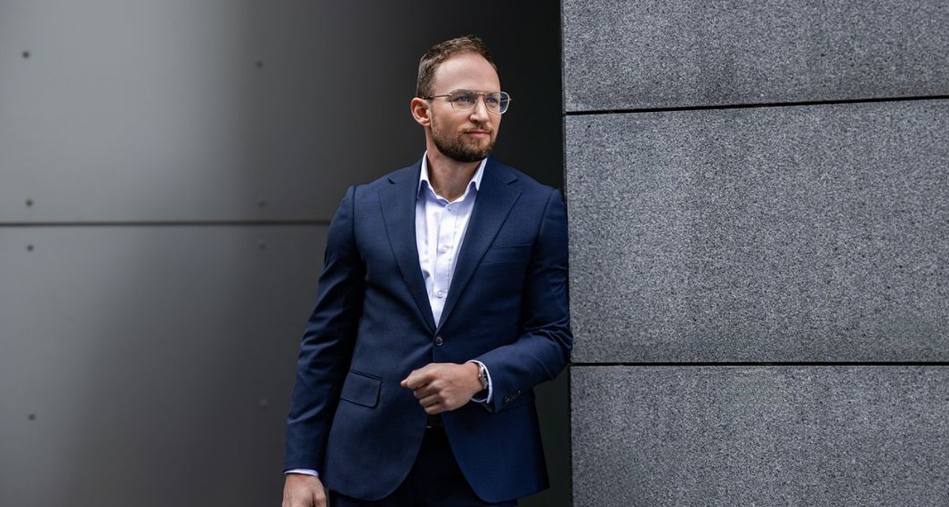 Portret mężczyzny w okularach i granatowym garniturze, opierającego się o szarą, teksturowaną ścianę, z zamyślonym spojrzeniem w bok.