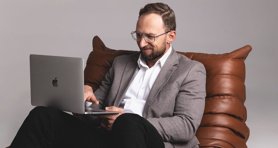 Uśmiechnięty mężczyzna w okularach i marynarce siedzi na skórzanym pufie i pracuje na laptopie Apple, patrząc na ekran.