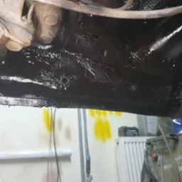 Podwozie samochodu po konserwacji antykorozyjnej, widoczne świeże, czarne zabezpieczenie, z elementami zawieszenia i śladami wcześniejszych napraw.