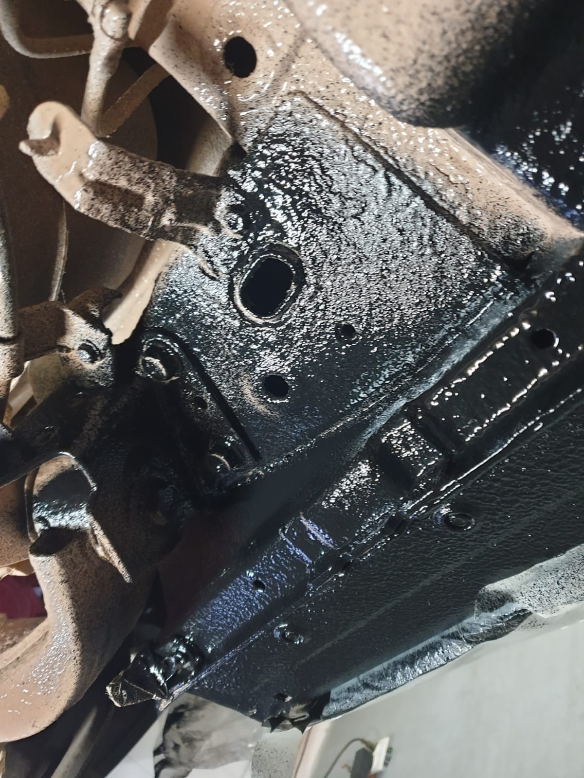 Podwozie pojazdu po nałożeniu grubej warstwy czarnego środka antykorozyjnego, widoczne elementy zawieszenia i liczne otwory technologiczne.