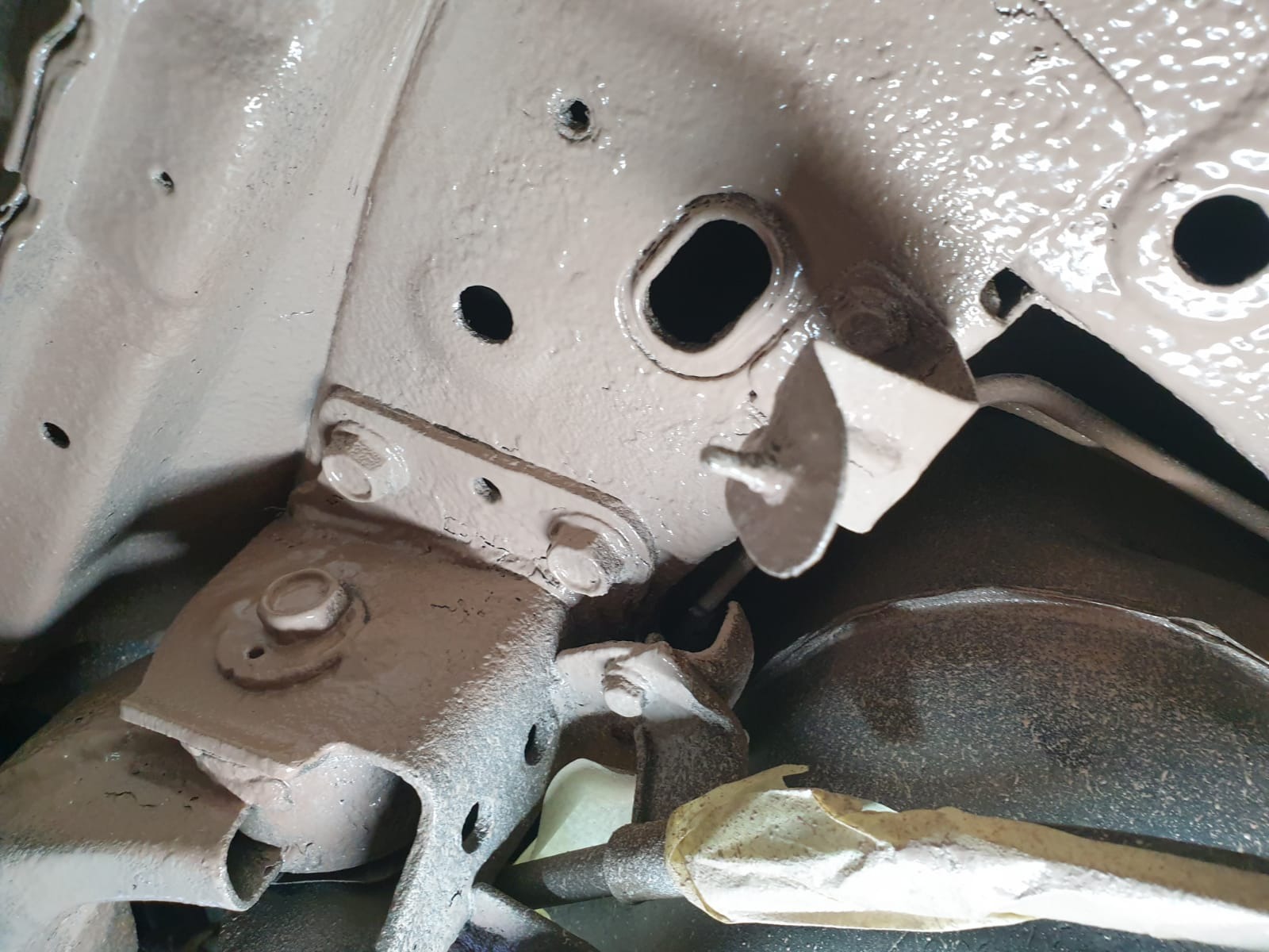 Podwozie samochodu po zabezpieczeniu antykorozyjnym, widoczne elementy zawieszenia pokryte jednolitą warstwą jasnobrązowej farby, fragment rury owiniętej taśmą maskującą.