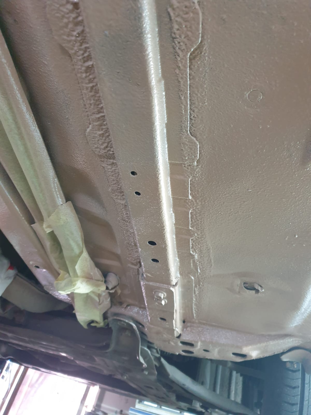 Podwozie samochodu po zabezpieczeniu antykorozyjnym, widoczne elementy metalowe pokryte jednolitą warstwą ochronną w kolorze beżowym, fragmenty zamaskowane taśmą malarską.