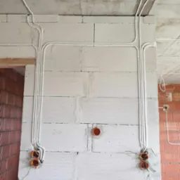 Białe kable elektryczne prowadzone po ścianie z białych bloczków betonu komórkowego, widoczne pomarańczowe puszki instalacyjne i surowy betonowy sufit.