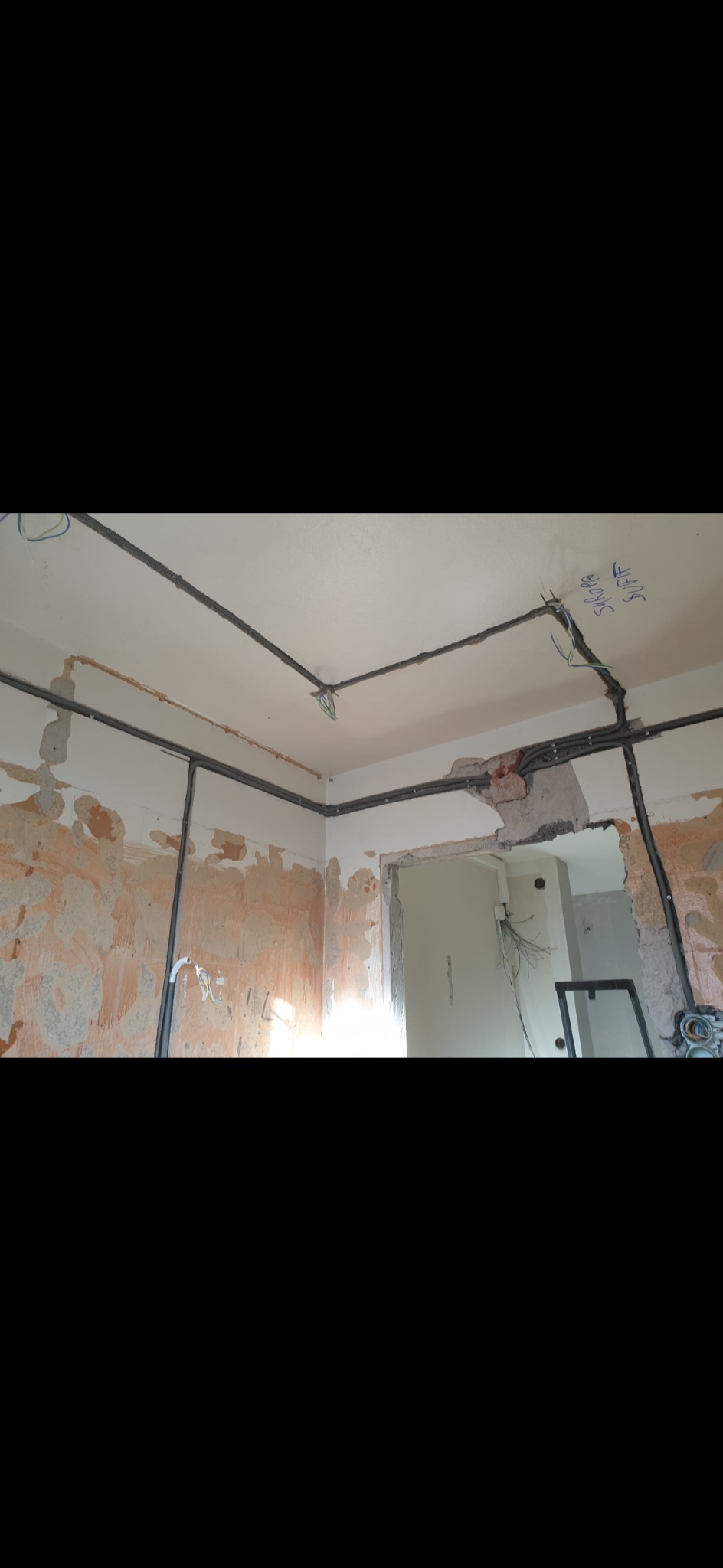 Szare korytka kablowe zamontowane na suficie i ścianach w surowym stanie, widoczne przewody elektryczne, ściany częściowo otynkowane, niebieskie napisy na suficie.