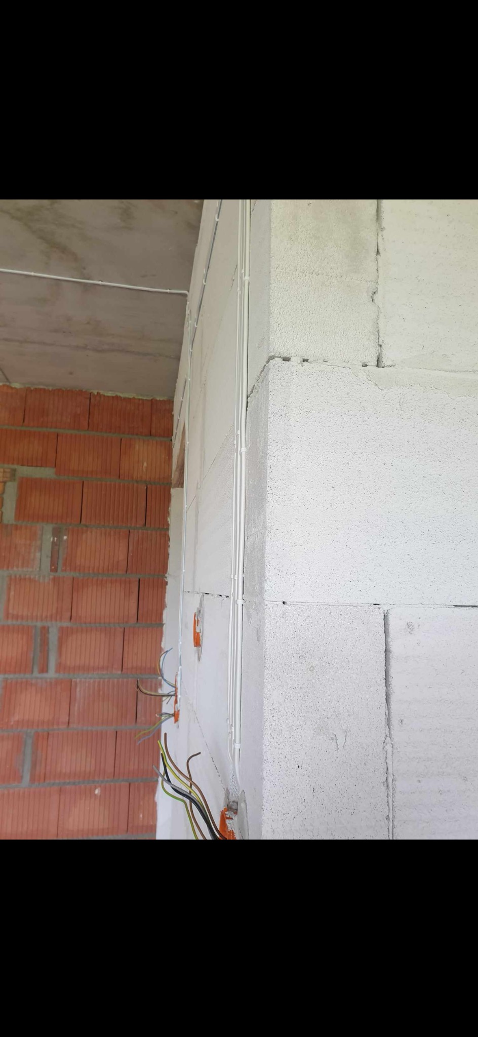 Surowy stan budynku: białe ściany z betonu komórkowego z poprowadzonymi przewodami elektrycznymi w białych rurkach, widoczne kable w puszkach instalacyjnych, ściana z czerwonej cegły w tle.