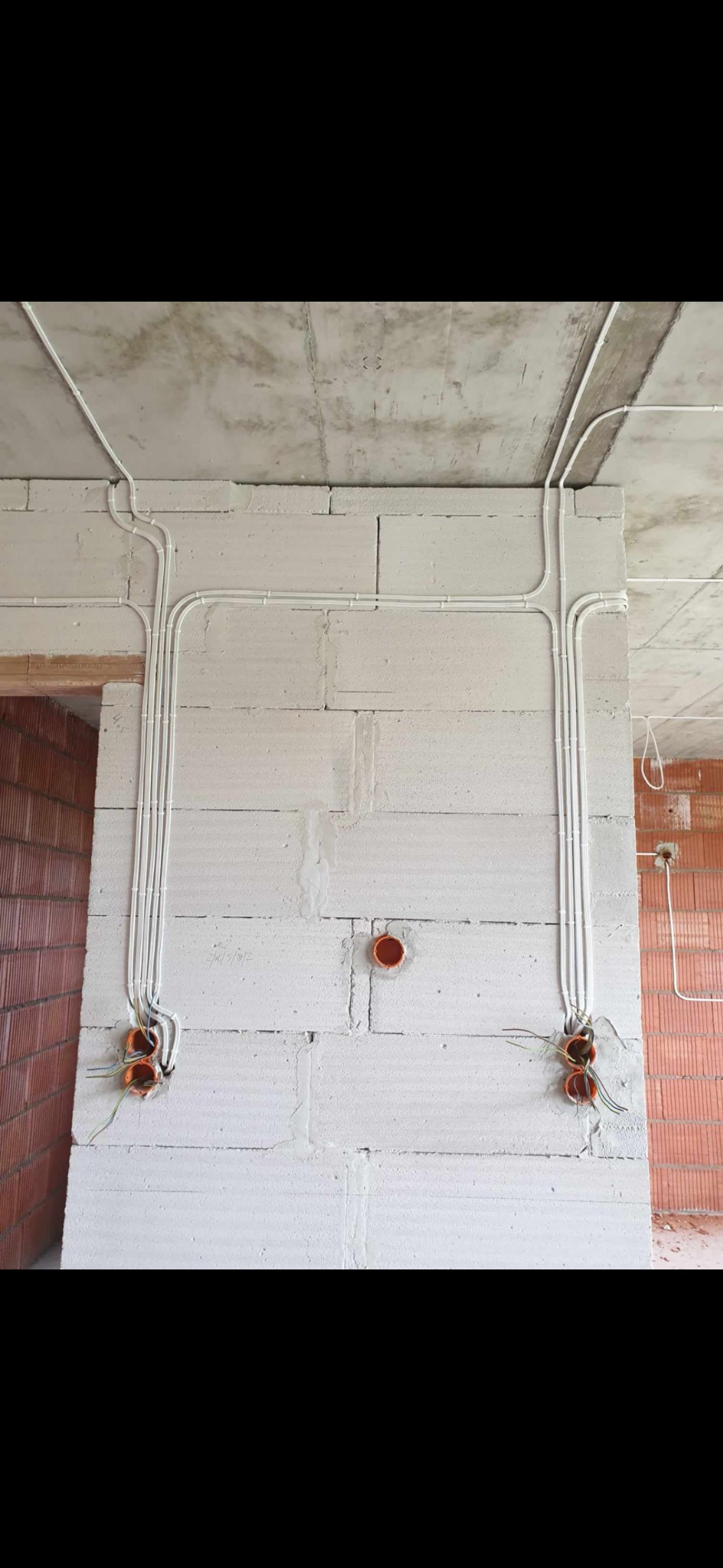 Białe kable elektryczne prowadzone po ścianie z białych bloczków betonu komórkowego, widoczne pomarańczowe puszki instalacyjne i surowy betonowy sufit.