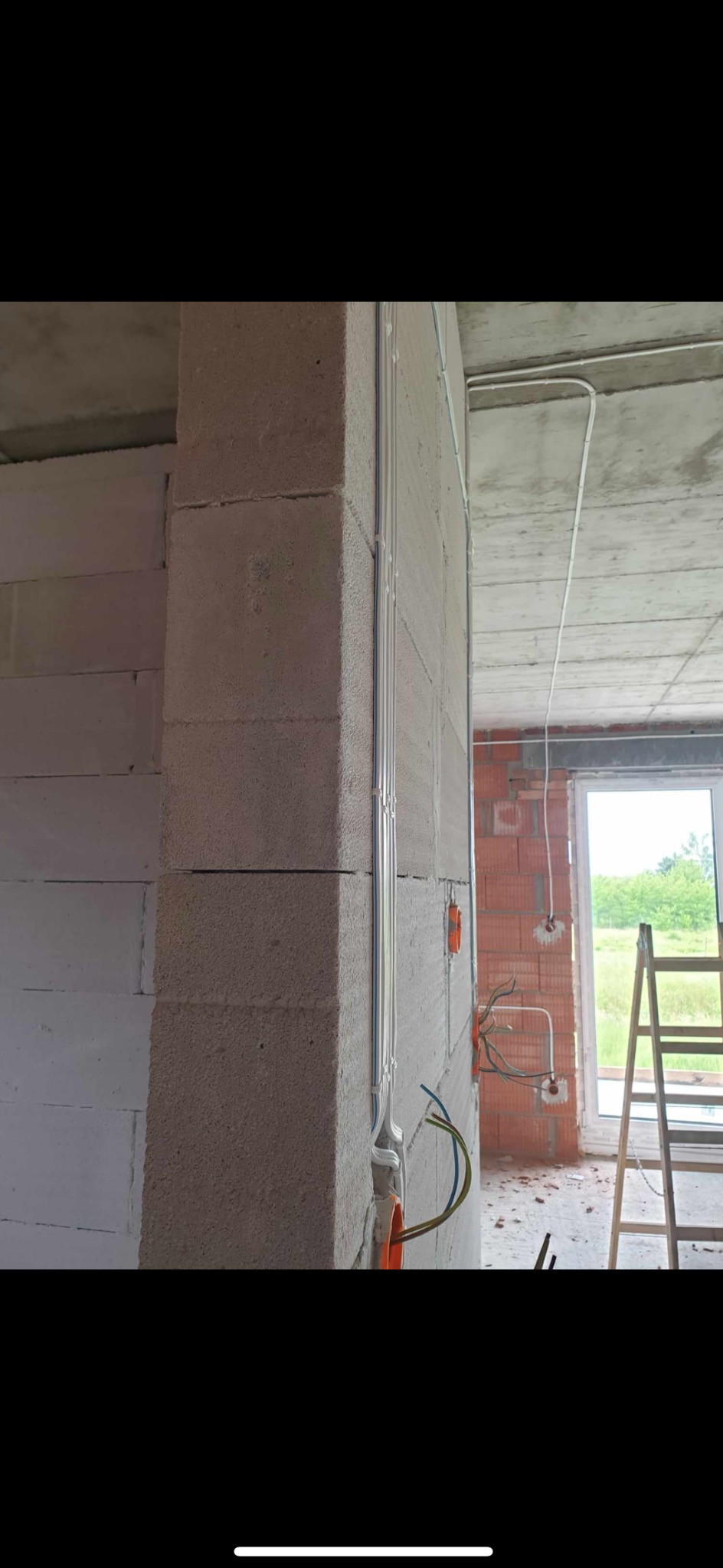 Surowy stan budynku w trakcie prac instalacyjnych: widoczne przewody elektryczne prowadzone po ścianie z bloczków betonowych, pomarańczowe puszki instalacyjne, drabina oparta o ścianę przy oknie.
