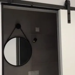 Przesuwne białe drzwi na czarnej szynie, odsłaniające fragment łazienki z szarymi płytkami, okrągłym lustrem na ścianie i rolką papieru toaletowego.