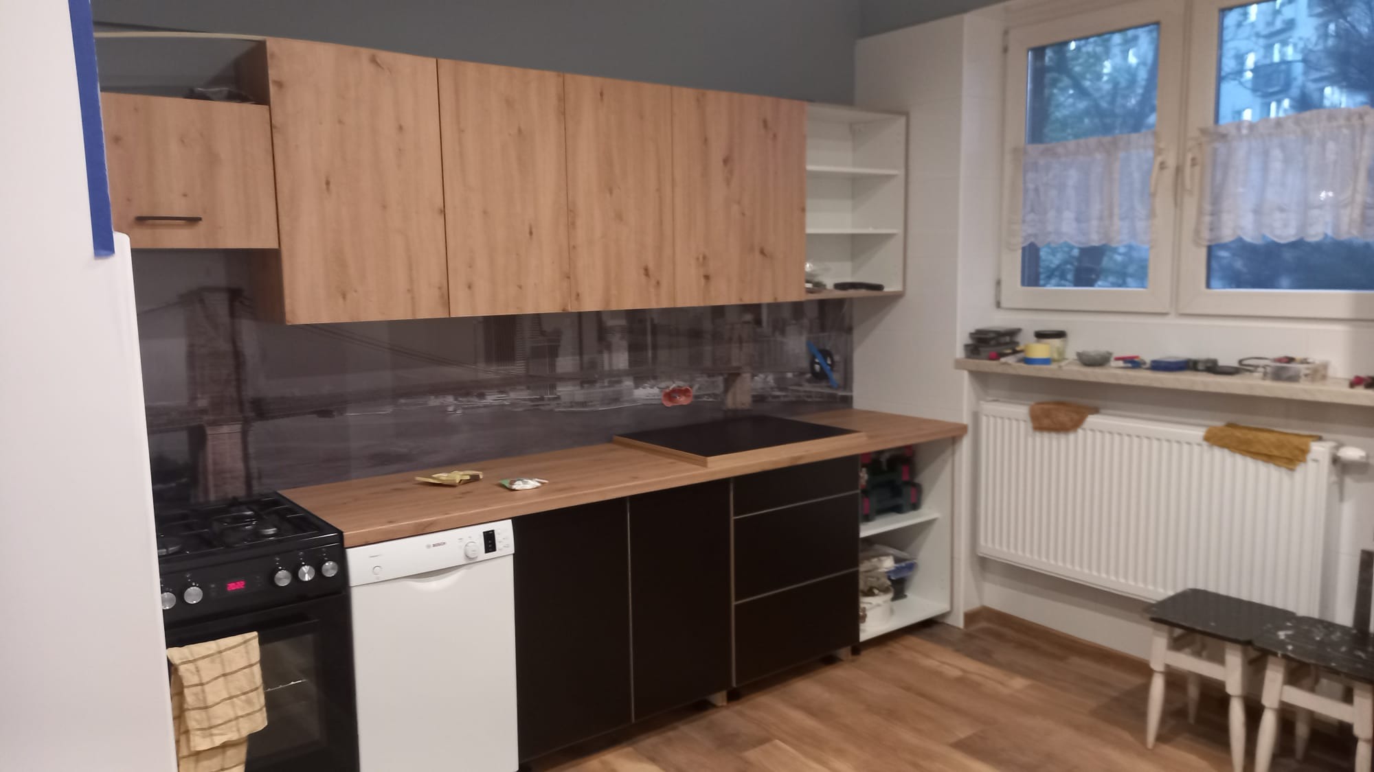 Kuchnia po remoncie: drewnopodobne szafki wiszące nad czarnymi szafkami dolnymi, zlew wpuszczony w blat, na ścianie fototapeta z motywem miejskim, okno z białymi firankami, grzejnik pod oknem.