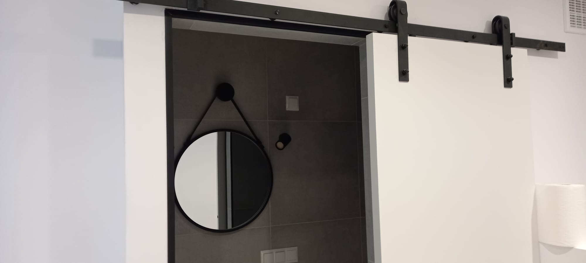 Przesuwne białe drzwi na czarnej szynie, odsłaniające fragment łazienki z szarymi płytkami, okrągłym lustrem na ścianie i rolką papieru toaletowego.
