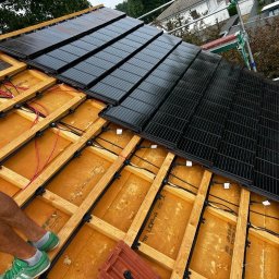 DLC Power Sp. z o.o. - Montaż paneli słonecznych na dachu domu, widoczne drewniane elementy konstrukcyjne, kable i fragment zielonych butów montera.