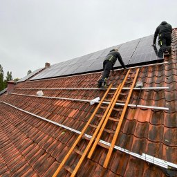 DLC Power Sp. z o.o. - Montaż paneli słonecznych na dachu z czerwonej dachówki, widoczni dwaj instalatorzy na drabinie i zamontowane szyny montażowe.