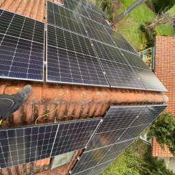 DLC Power Sp. z o.o. - Montaż paneli słonecznych na dachu z czerwonej dachówki, widoczny fragment buta montera i komin na tle zieleni ogrodu.