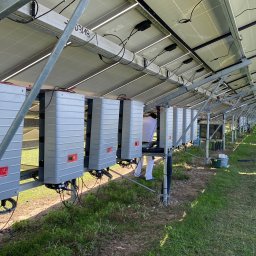 DLC Power Sp. z o.o. - Rząd szarych falowników zamontowanych pod panelami słonecznymi na metalowej konstrukcji, widoczny pracownik w białym kombinezonie podczas instalacji, w tle trawa i kolejne rzędy paneli.