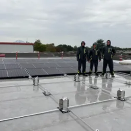 Trzech monterów w odzieży roboczej stoi na dachu z częściowo zamontowanymi panelami słonecznymi i aluminiową konstrukcją wsporczą, widok na budynek z logo BAAS w tle, pochmurne niebo.