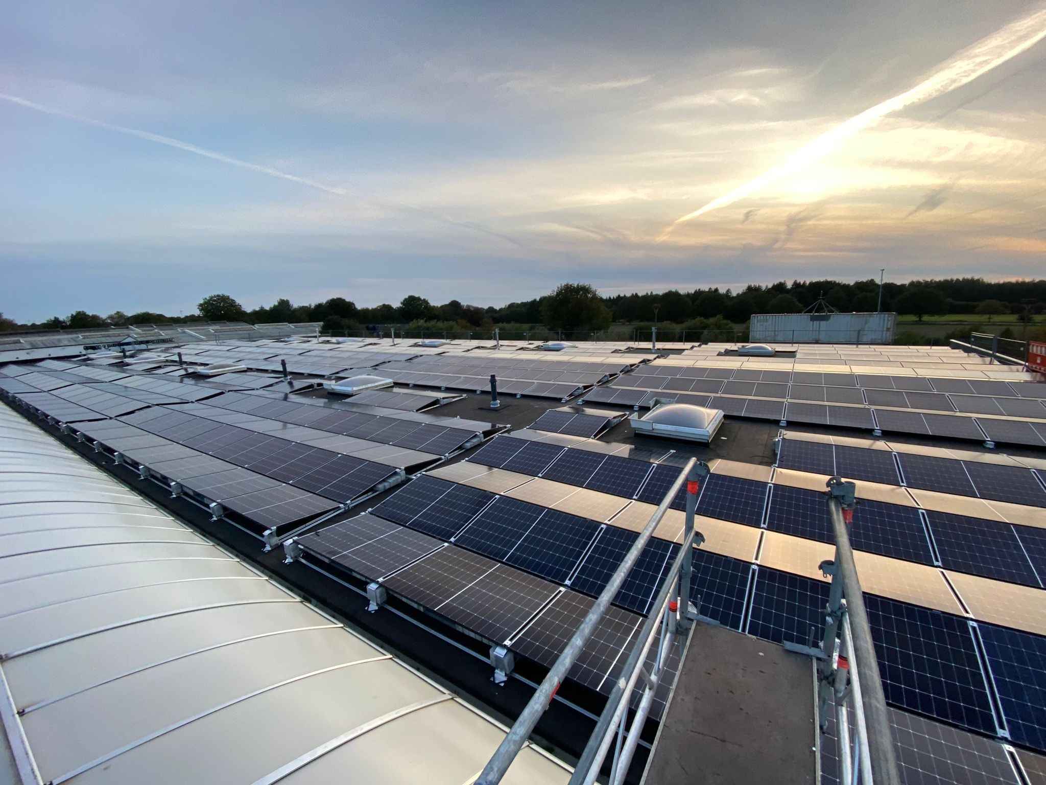 Widok z góry na rozległą instalację paneli słonecznych na dachu budynku przemysłowego, z widocznym rusztowaniem i fragmentem nieba z chmurami.