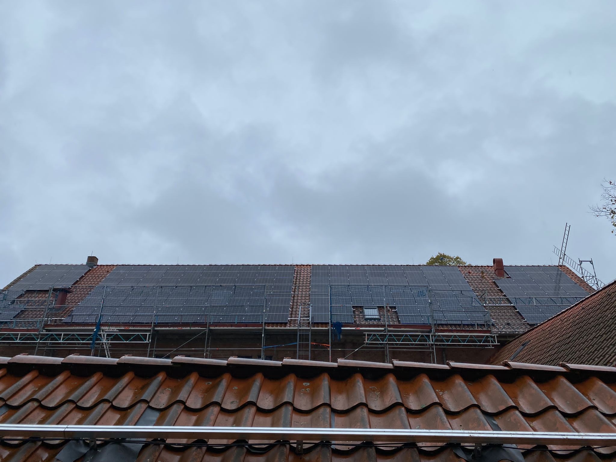 Montaż paneli fotowoltaicznych na zabytkowym budynku z czerwonej cegły, widoczne rusztowania i pochmurne niebo.