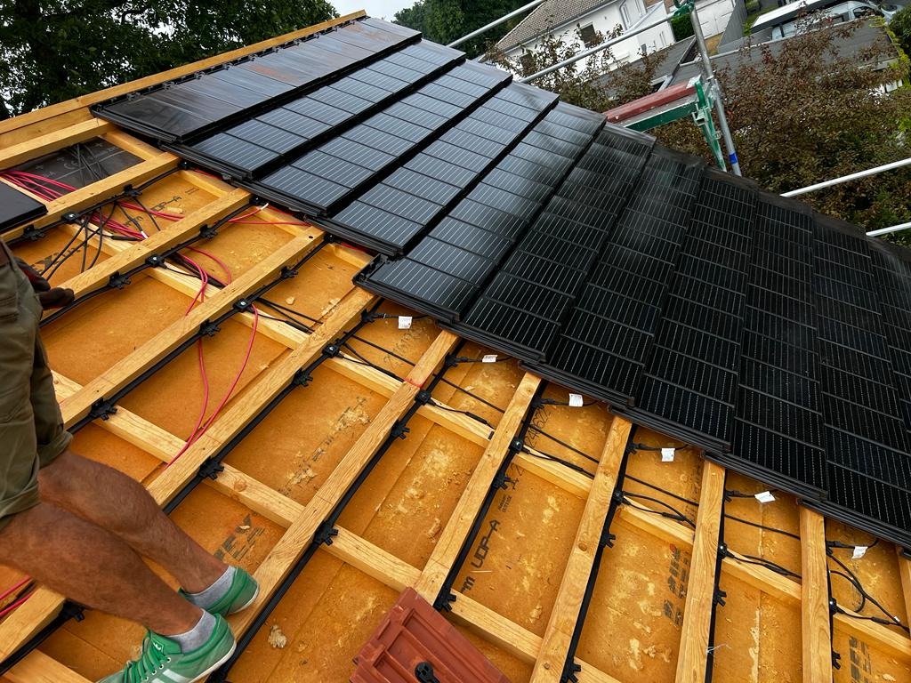 Montaż paneli słonecznych na dachu domu, widoczne drewniane elementy konstrukcyjne, kable i fragment zielonych butów montera.