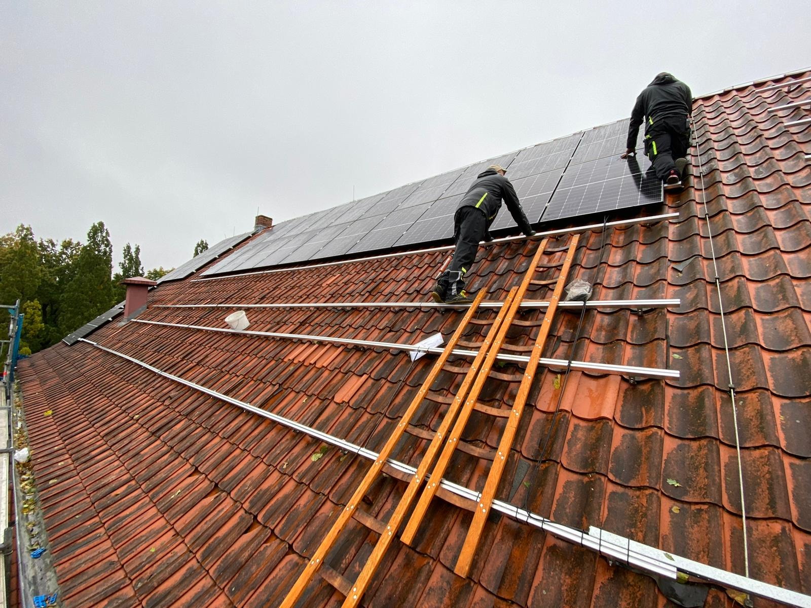 Montaż paneli słonecznych na dachu z czerwonej dachówki, widoczni dwaj instalatorzy na drabinie i zamontowane szyny montażowe.