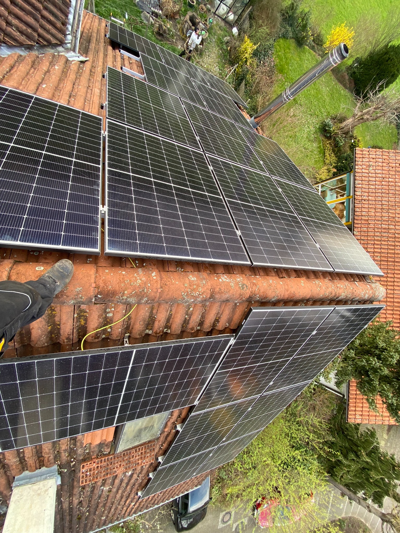 Montaż paneli słonecznych na dachu z czerwonej dachówki, widoczny fragment buta montera i komin na tle zieleni ogrodu.