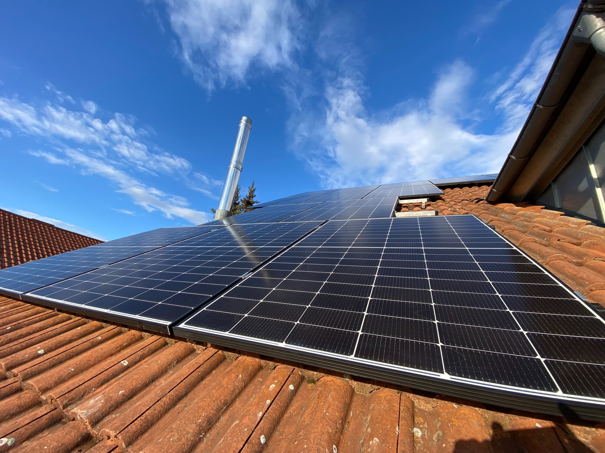 Panele słoneczne na dachu z czerwonej dachówki, widok z dołu, błękitne niebo z chmurami w tle, komin wentylacyjny obok.