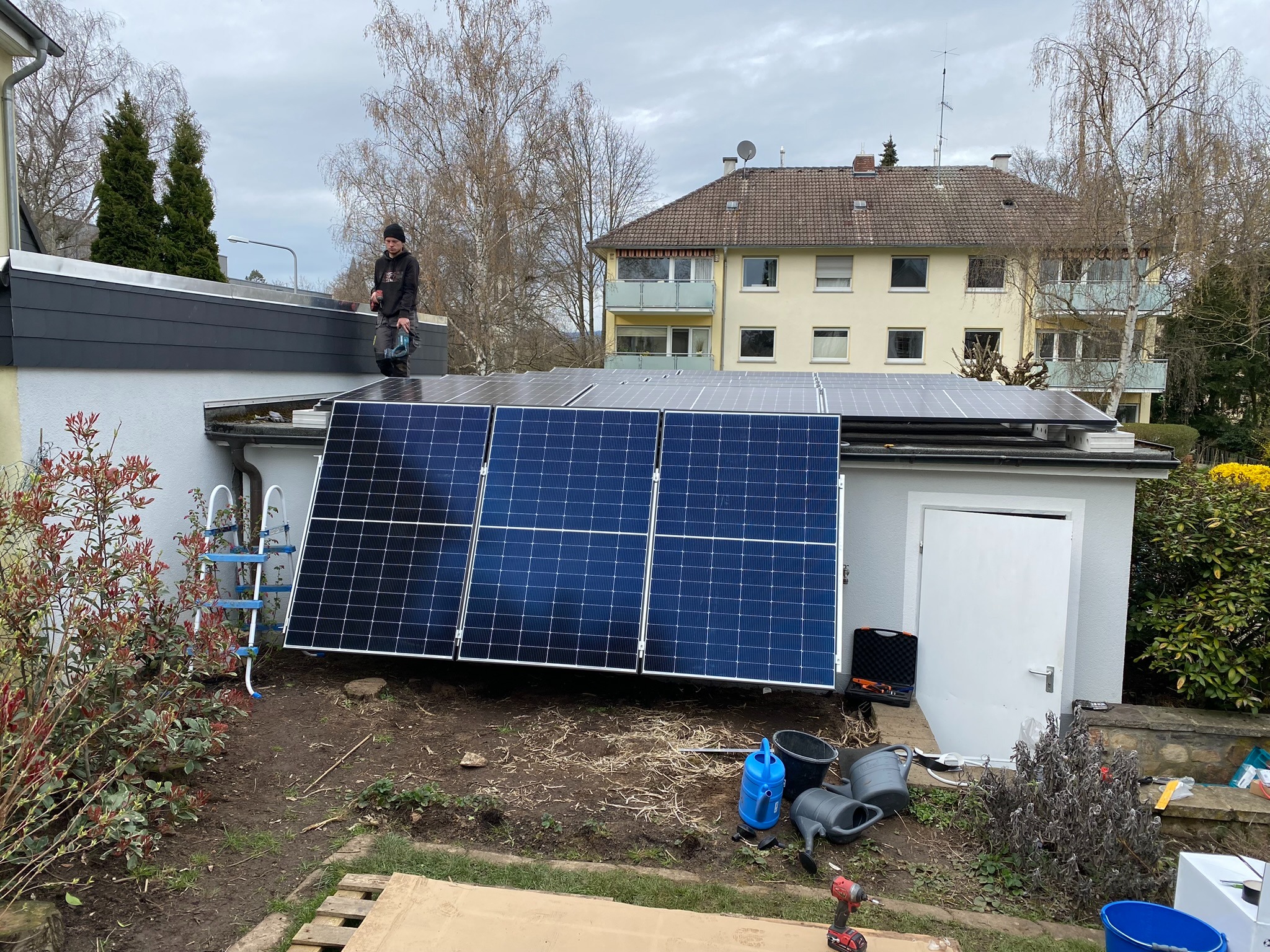 Instalator z wiertarką na dachu budynku podczas montażu paneli słonecznych; w tle dom mieszkalny, drabina oparta o ścianę, narzędzia i akcesoria na ziemi.