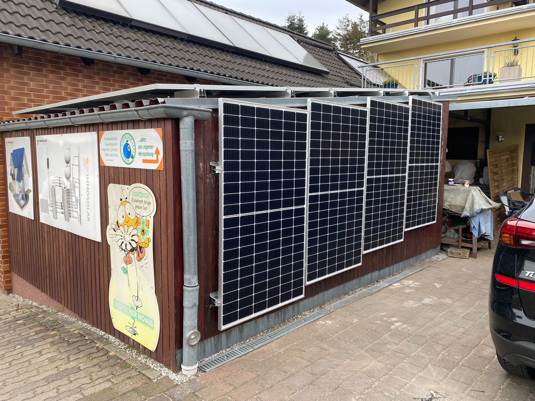 Panele słoneczne zamontowane na drewnianej konstrukcji przy budynku, widoczne reklamy na ścianie i fragment samochodu.