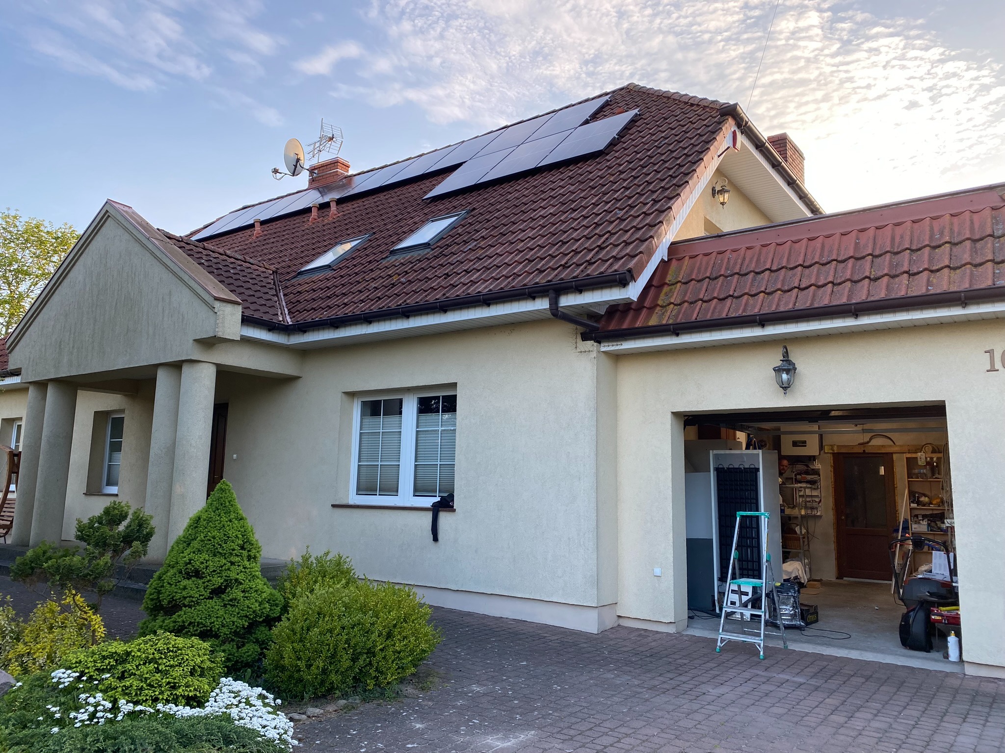 Dom jednorodzinny w Lipianach z panelami fotowoltaicznymi na czerwonym dachu i anteną satelitarną, widok z zewnątrz garażu z modułami fotowoltaicznymi i drabiną.