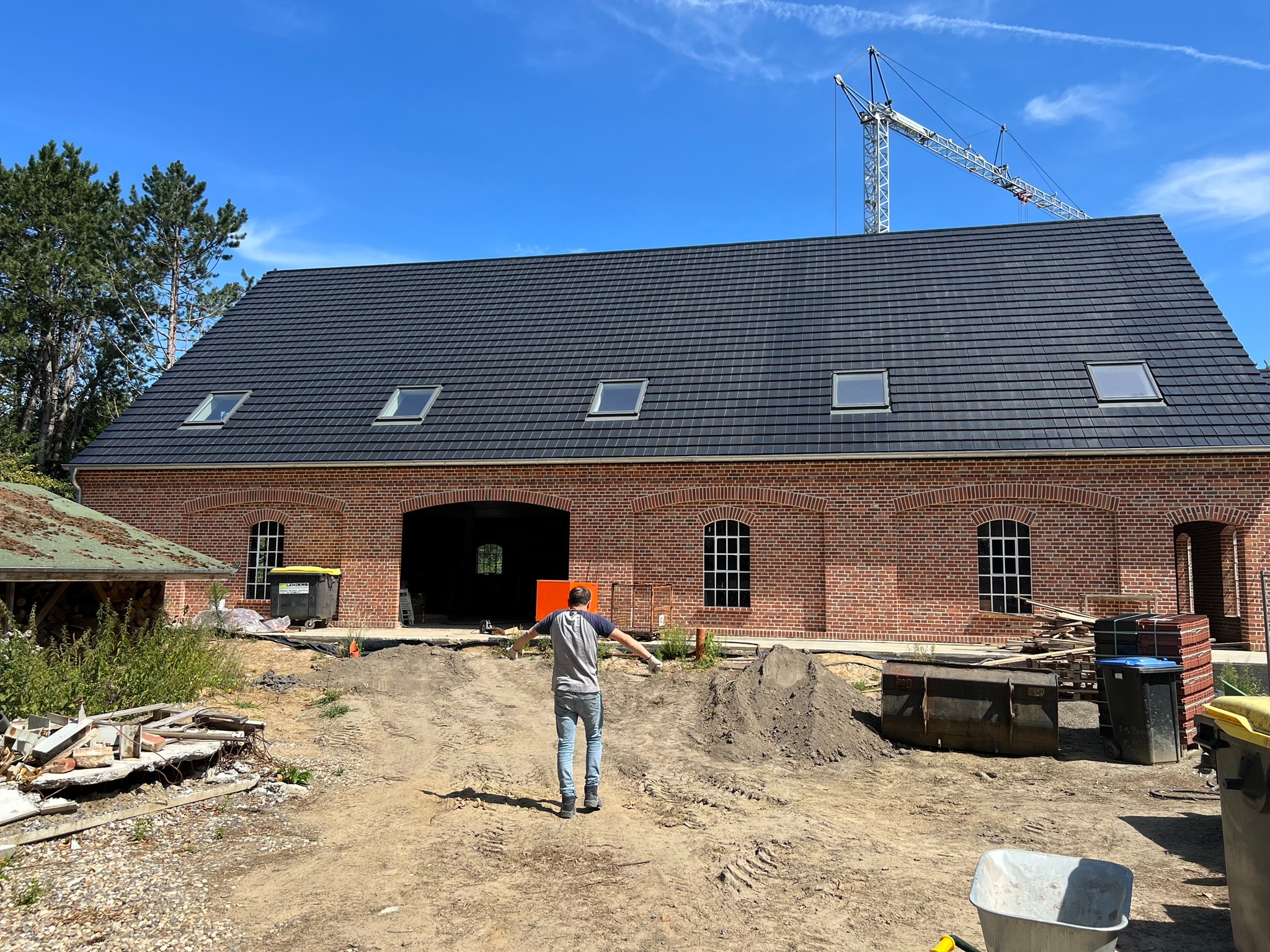 Nowy budynek z czerwonej cegły z ciemną, skośną dachówką i oknami dachowymi, widok z tyłu mężczyzny stojącego na placu budowy z rozłożonymi rękami, w tle żuraw budowlany.