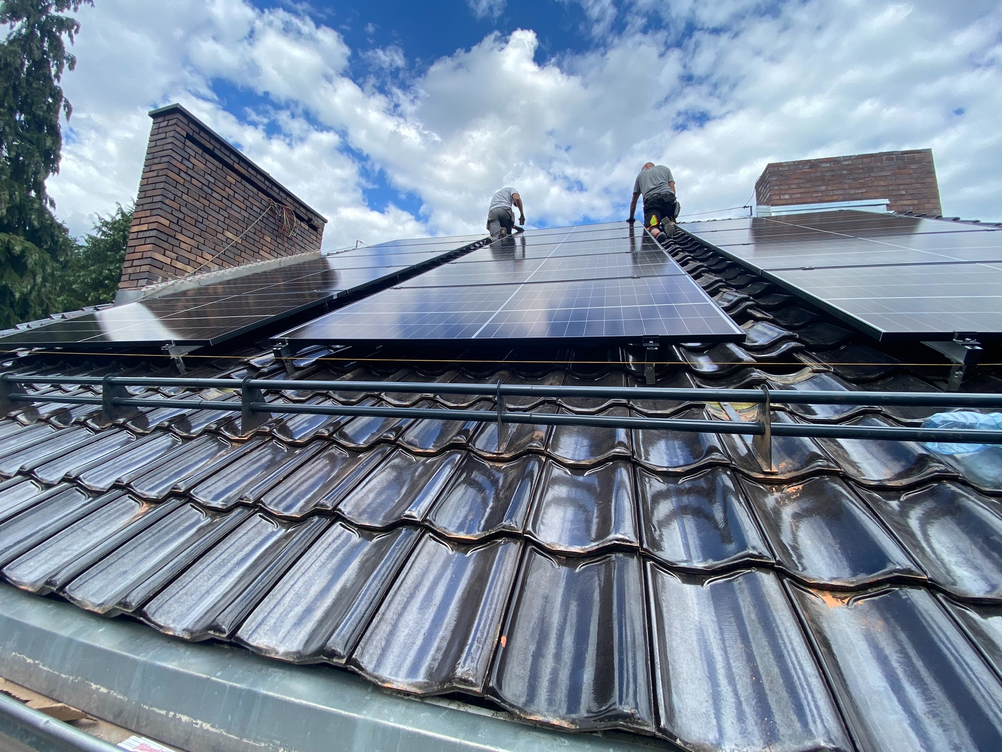 Montaż paneli słonecznych na dachu z ciemnej dachówki ceramicznej; dwóch monterów pracuje na wysokości, widoczne kominy i błękitne niebo z chmurami.